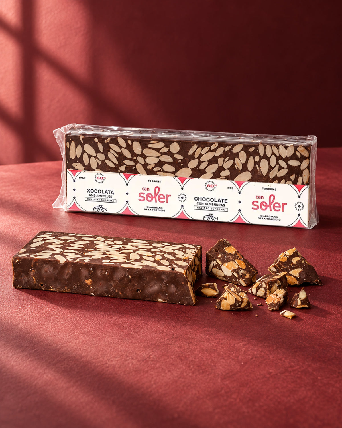 Turrón de chocolate 60 % con almendras de Can Soler en su envase, con una barra sin envase y trozos partidos delante