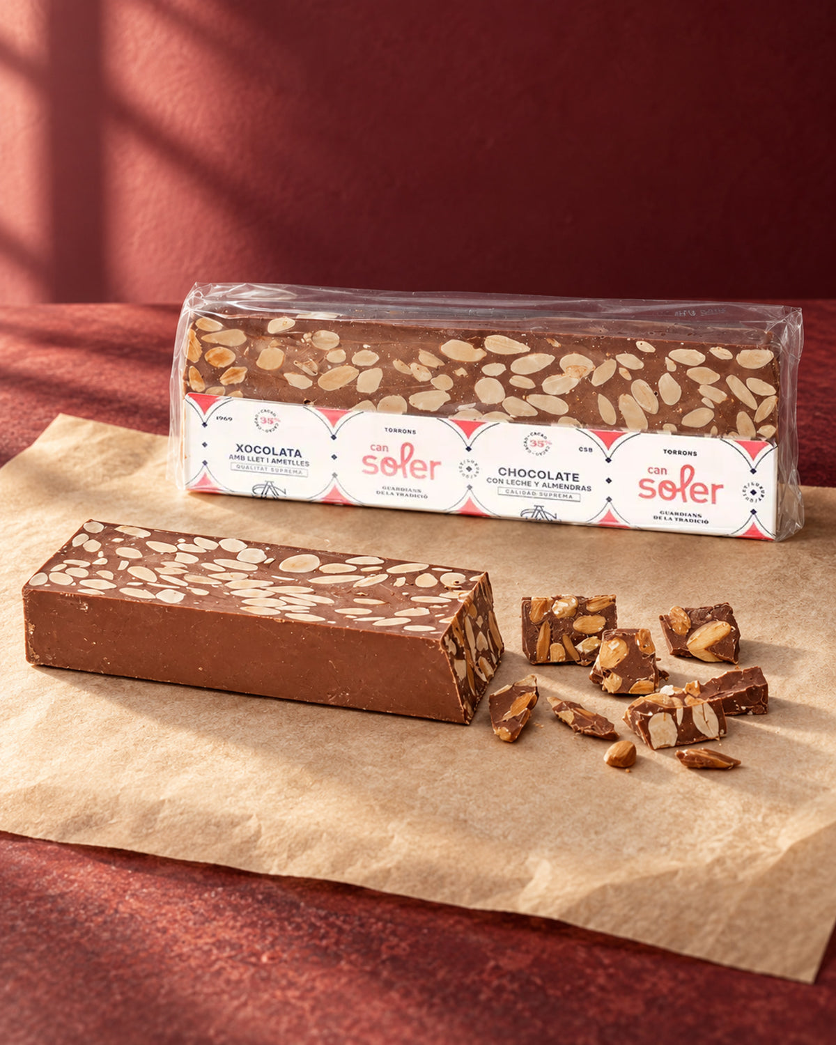 Turrón de chocolate con leche y almendras de Can Soler: barra sin envase y trozos partidos delante del paquete con etiqueta