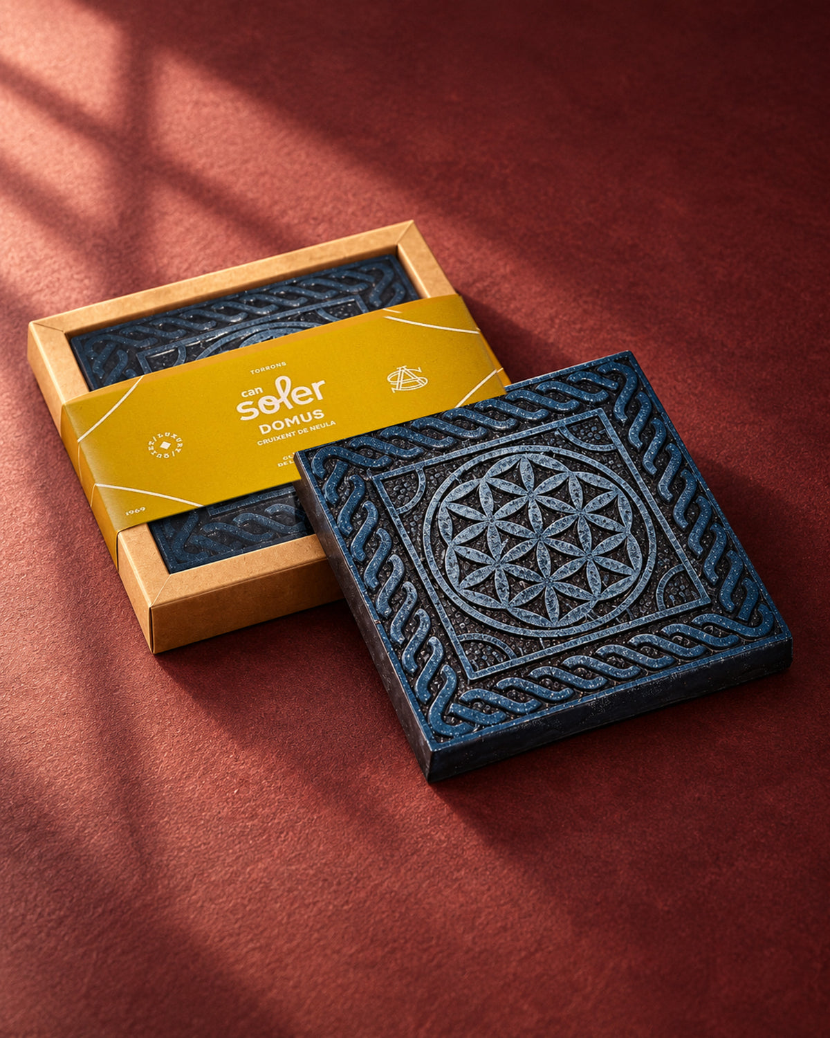 Chocolate de Museo «Domus» de Can Soler: placa con relieve geométrico de mosaico y orla trenzada junto a su estuche con faja mostaza