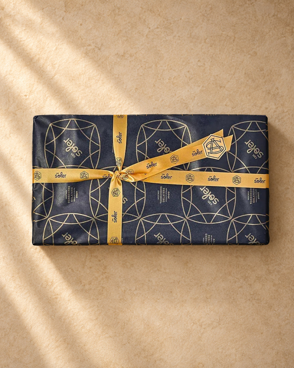 Vista desde arriba del lote de regalo «Para quedar bien» de Can Soler, envuelto en papel oscuro estampado y lazo dorado de la marca