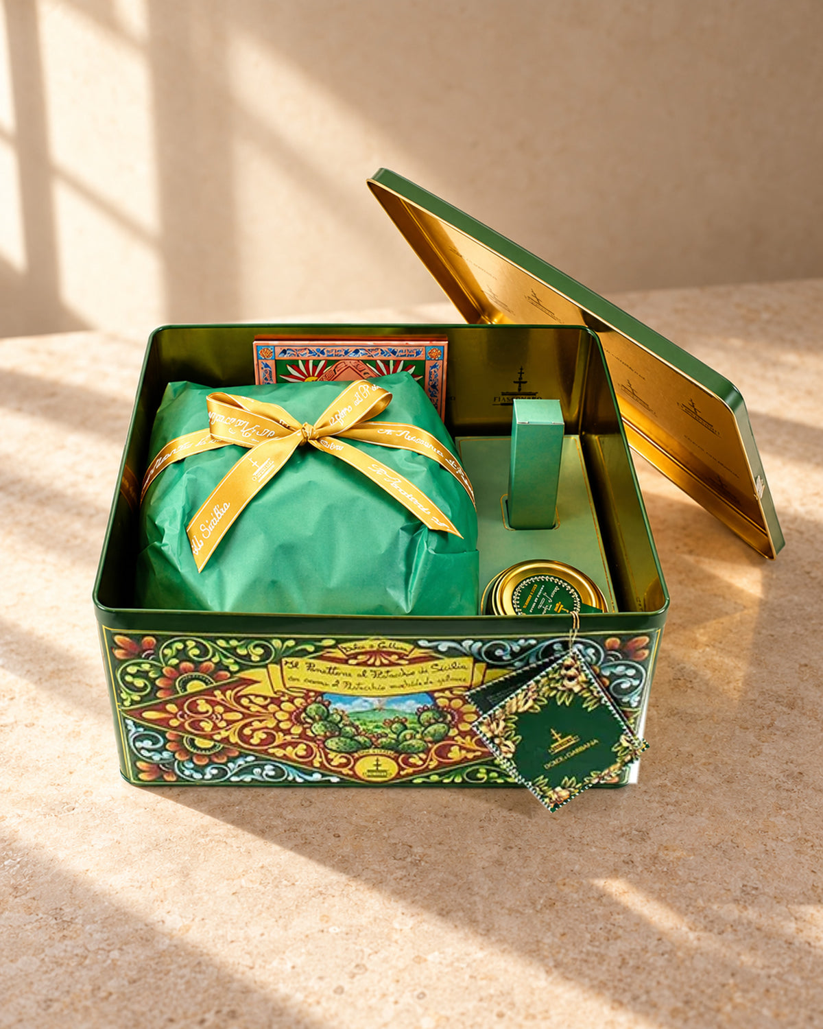 Lata Dolce&Gabbana abierta con el panettone Fiasconaro de pistacho envuelto en papel verde con lazo dorado y un tarrito