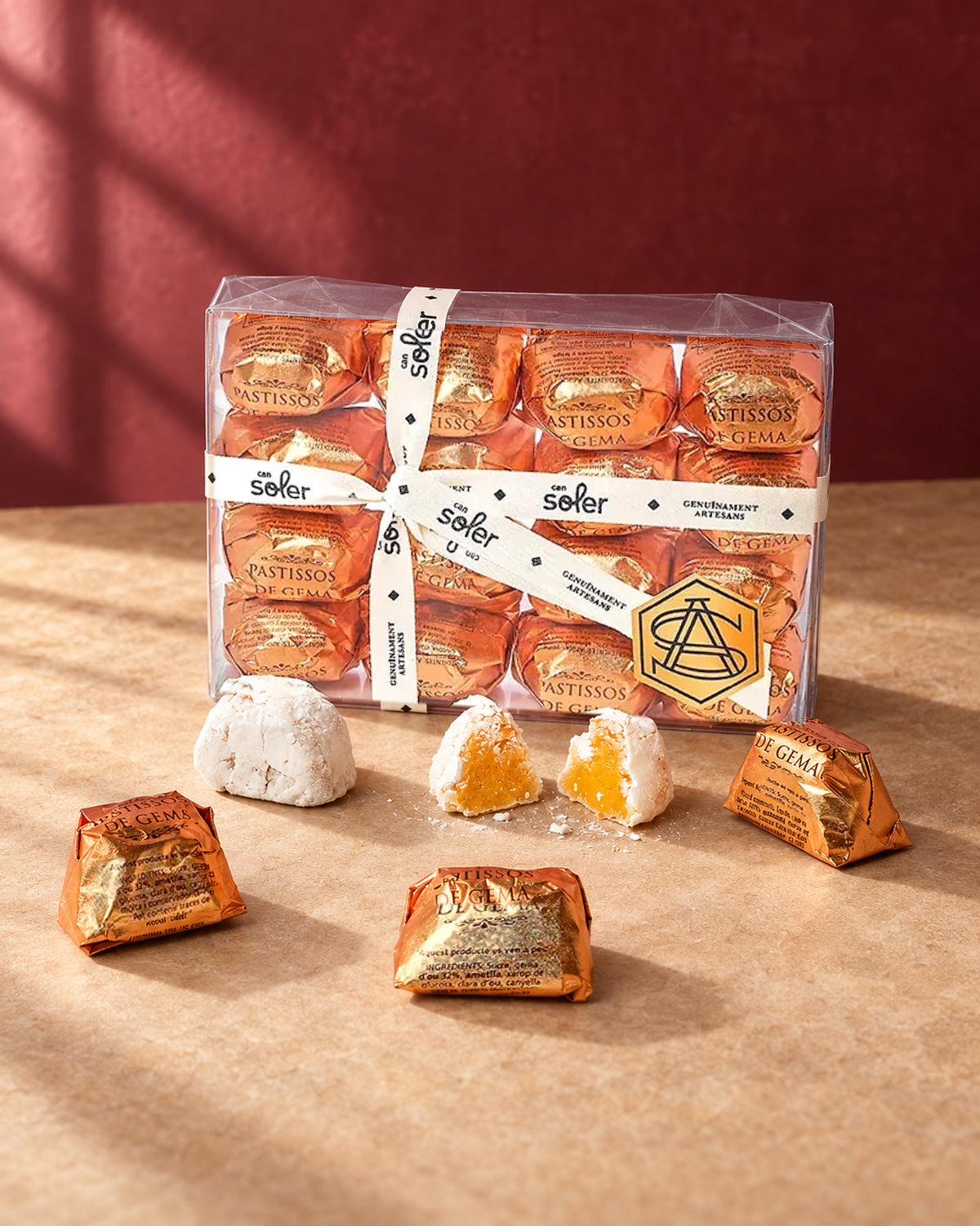 Caja transparente de pastelitos de mazapán de yema Can Soler con cinta, piezas envueltas en dorado y una partida con su relleno