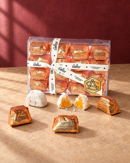 Caja transparente de pastelitos de mazapán de yema Can Soler con cinta, piezas envueltas en dorado y una partida con su relleno